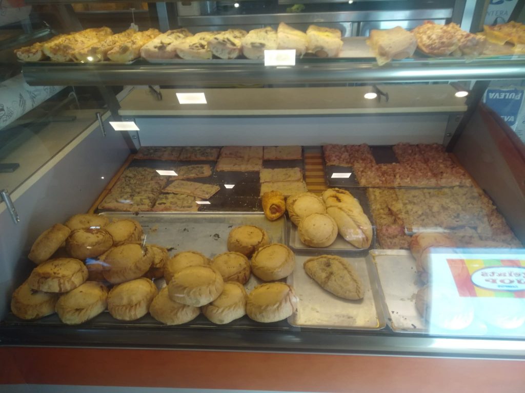 Imagen del mostrador de comida donde pueden verse empanadas, pizza, cocarrois, pan pizza, etc.